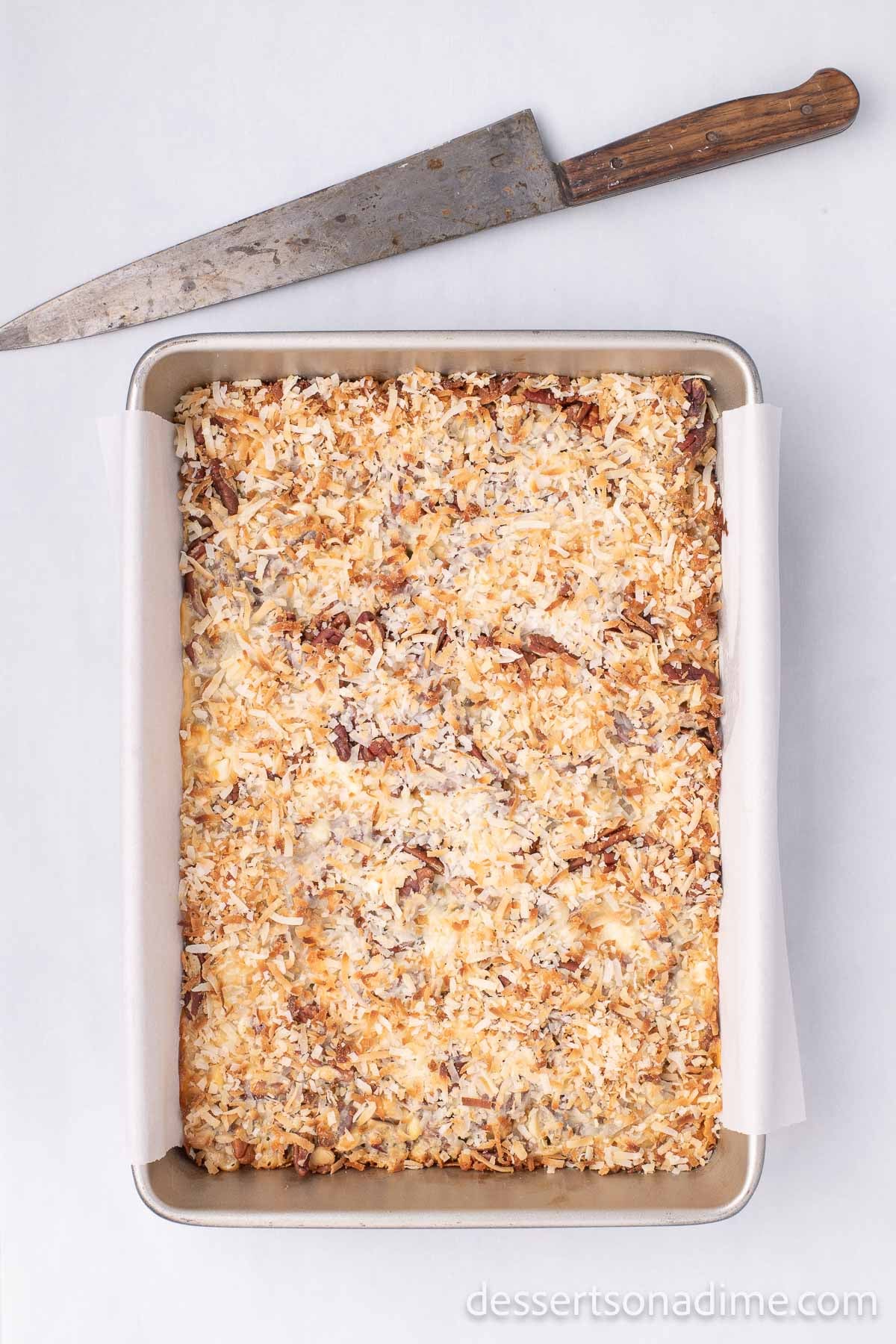 The 7 Layer bars cooling completely in the baking dish with a knife above the pan ready to cut the bars to be able to enjoy them! 