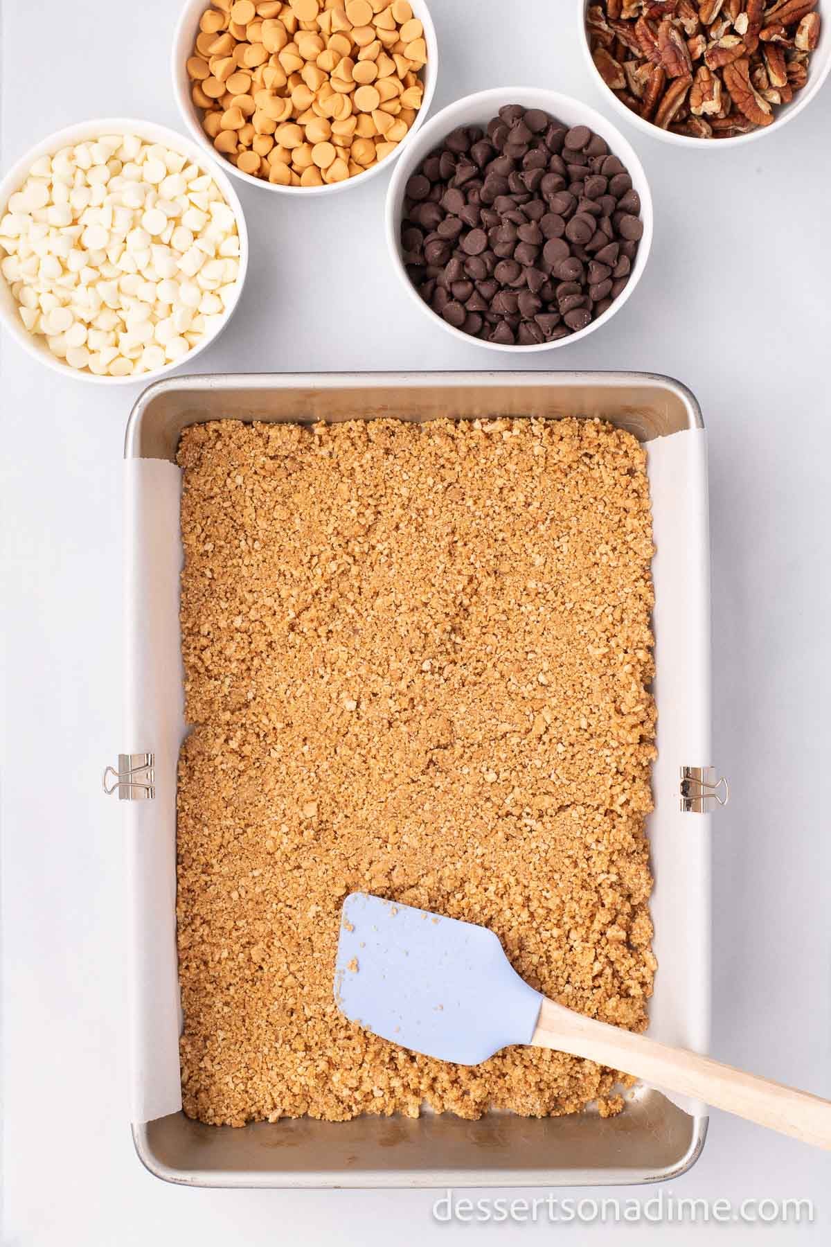 The graham cracker crumb mixture being pressed down into the baking dish lined with parchment paper.  
