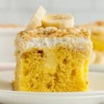 Side view of a slice of this banana pudding poke cake on a white plate topped with crushed vanilla wafers and sliced bananas