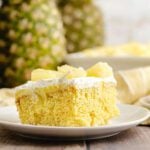 A piece of the pineapple poke cake on a white plate with diced up pineapple on top of the cake and 2 whole pineapples behind it.