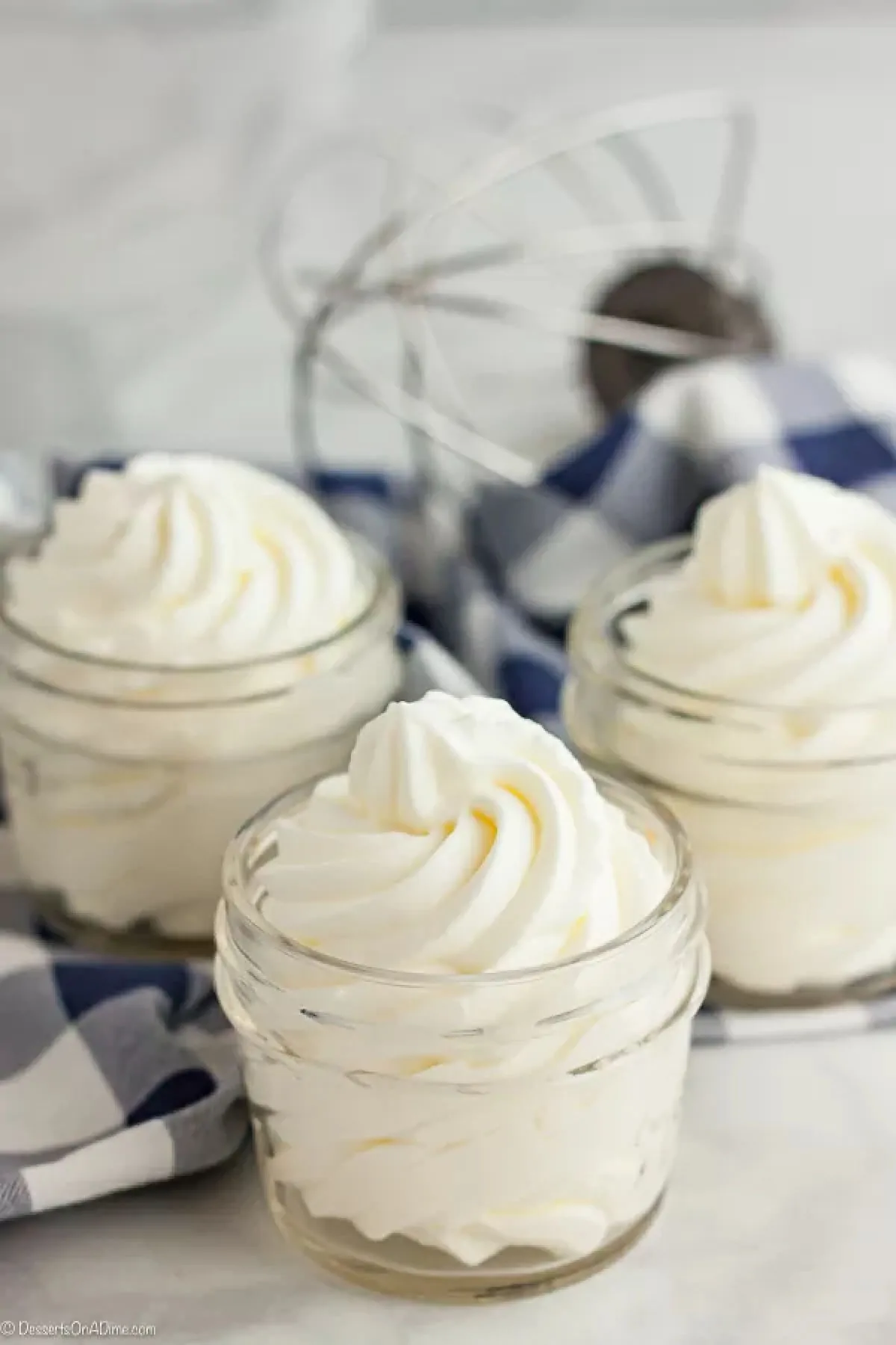 Once you try this Homemade Whipped Cream Recipe, you will never buy store bought whipped cream. This whipped cream recipes is easy with just 2 ingredients and tastes amazing. Try this heavy whipping cream recipes today. You will love this fresh whipped cream recipe! #dessertsonadime #whippedcream Homemade whipped cream in a small mason jar with 2 other jars and a whisk attachment behind it.