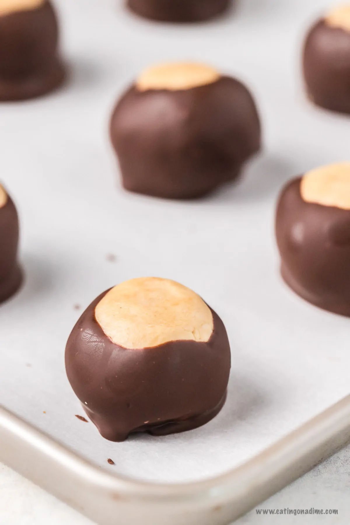 This no bake Buckeye balls recipe will be a hit for chocolate and peanut butter fans. Buckeye balls taste like peanut butter cups and melt in your mouth. This buckeye balls easy recipe is great for Christmas and no shortening required. #dessertonadime #buckeyeballsrecipe #buckeyeballsbest Buckeye Peanut Butter Balls on a parchment paper lined cookie sheet.
