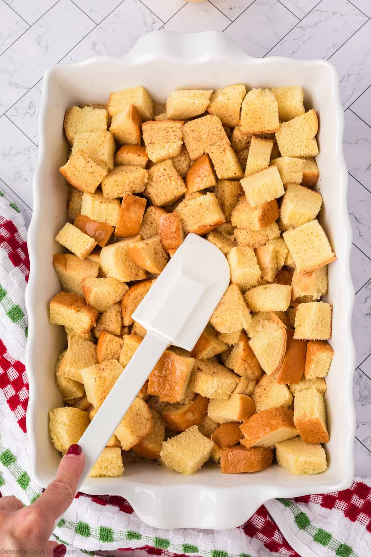 Cubed bread in casserole dish with spatula pressing mixture down. 