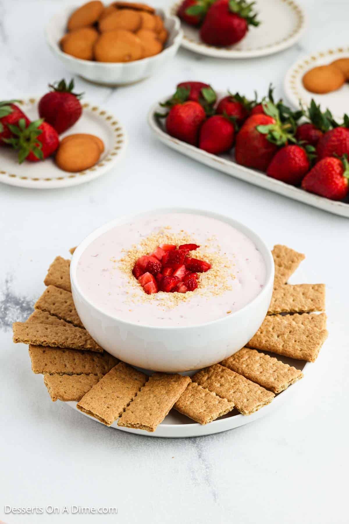 Bowl of Strawberry Cheesecake Dip topped with diced strawberries on a tray with graham crackers. 