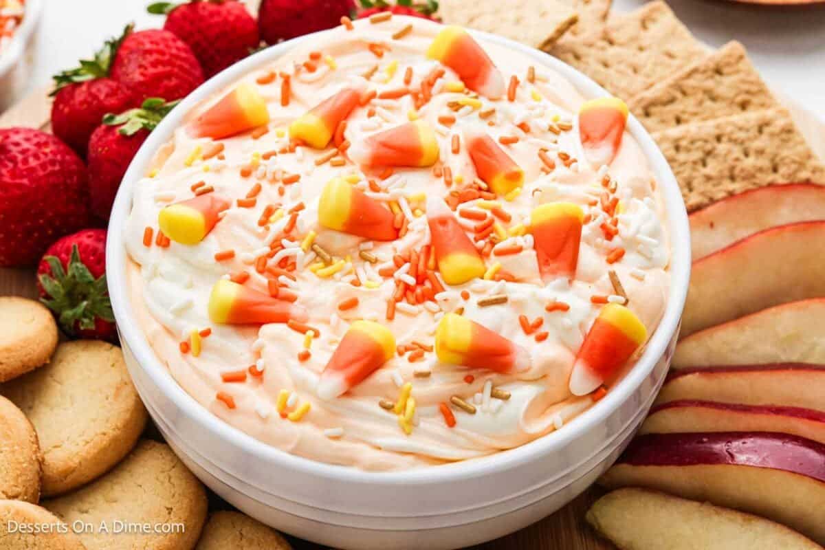 Candy Corn Dip in a white bowl topped with candy corn and sprinkles surrounded by fruit and cookies.