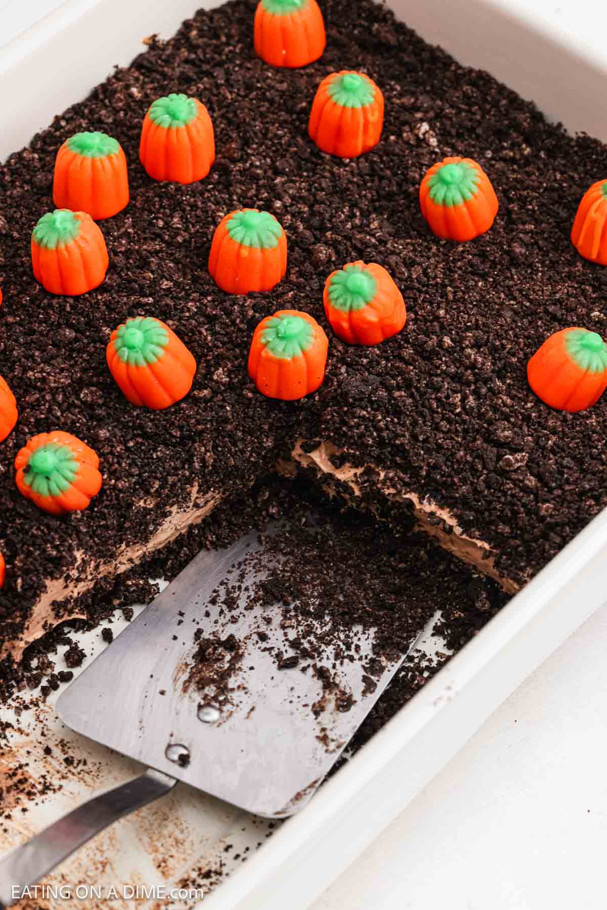 No bake dessert in a baking dish with crushed cookies and candy pumpkins on top being served with a spatula. 